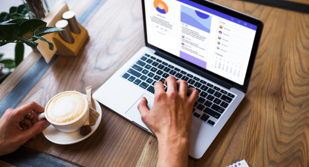 Na obrázku je počítač na stole. Vedle něj, na levé straně, je šálek kávy a dvě ruce.